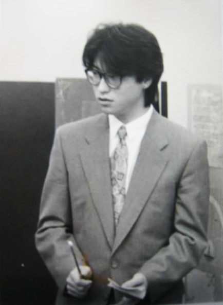 モノクロ写真。大きな黒縁メガネをかけ、ネクタイを締めた、大学院時代の熊澤先生